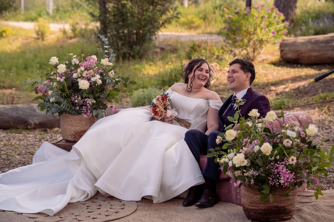 Novios sonriendo sentados juntos entre arreglos florales durante su boda al aire libre