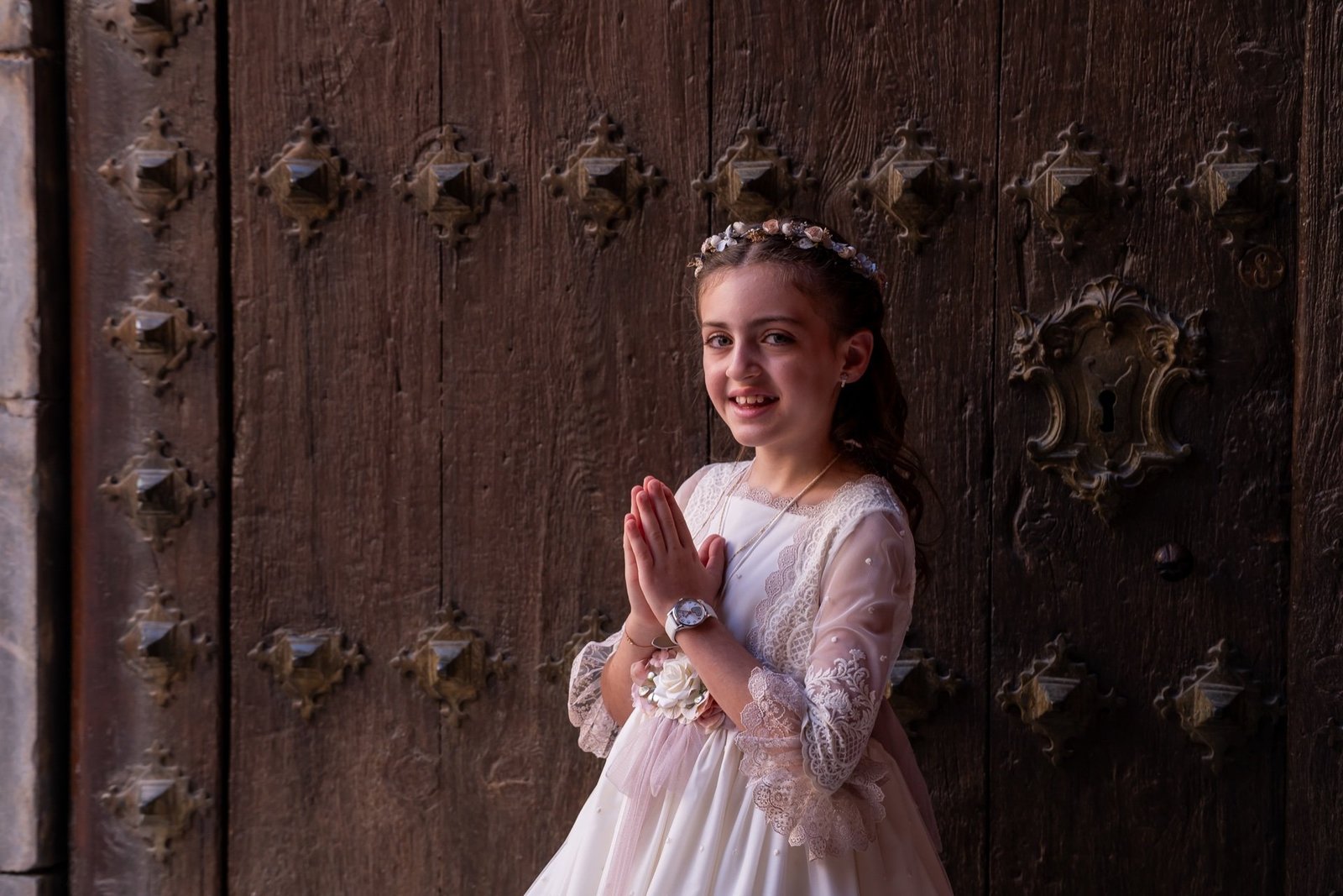Niña con vestido de comunión y las manos juntas delante de una puerta de madera ornamentada.