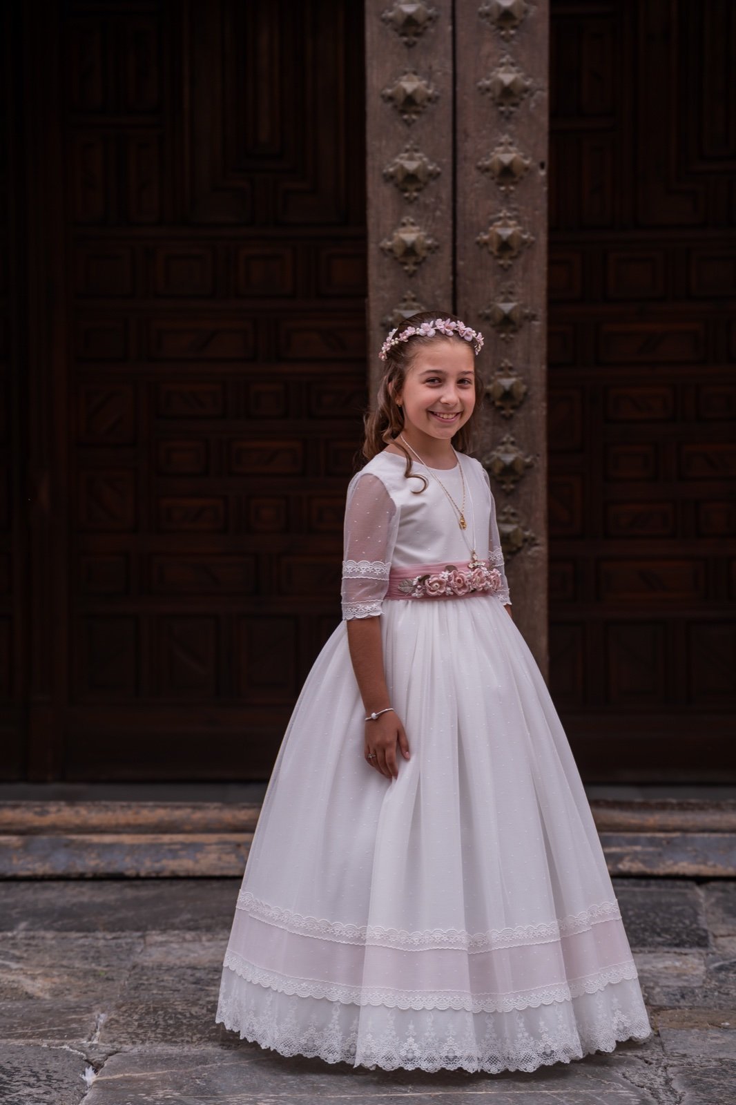 Niña con vestido de comunión y fajín rosa ante las puertas de la iglesia