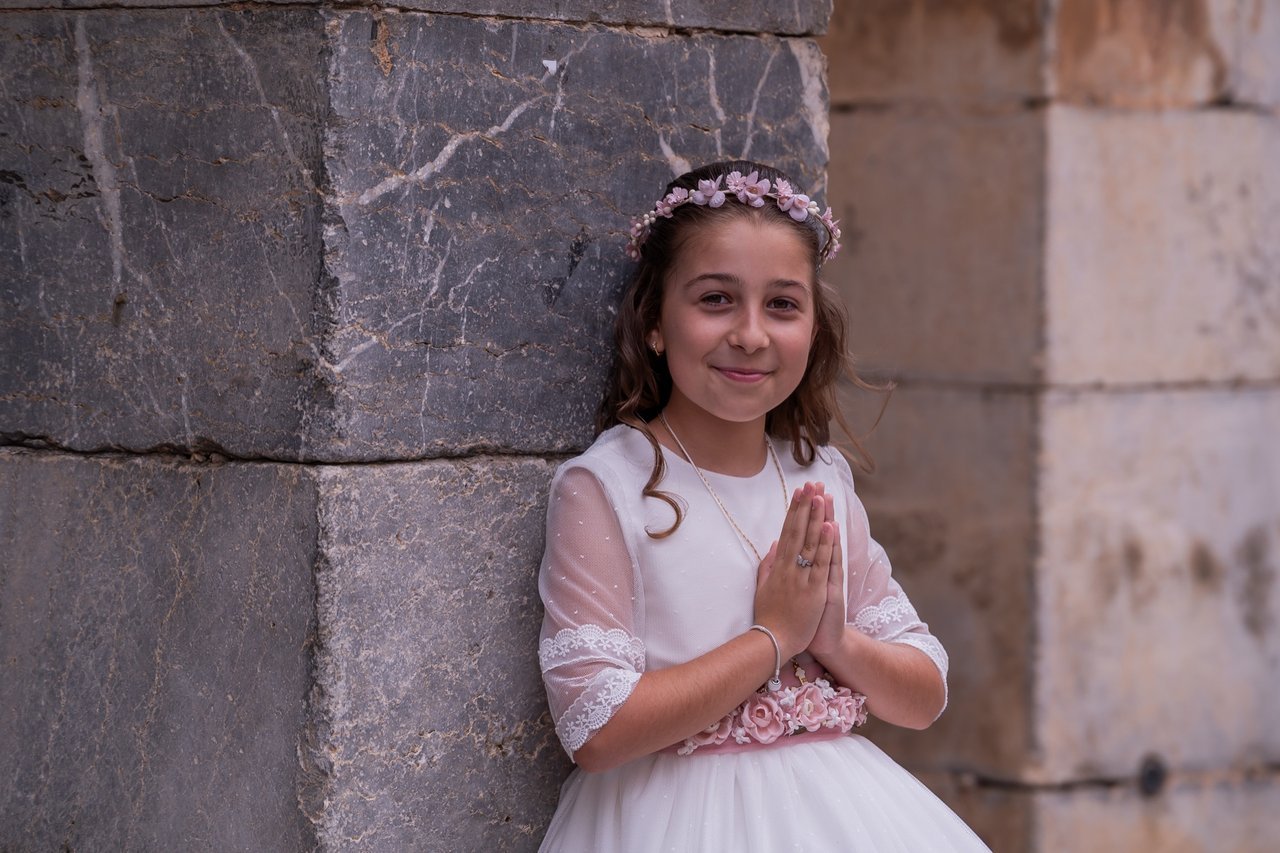 Niña con vestido de comunión y las manos juntas junto a un muro de piedra.