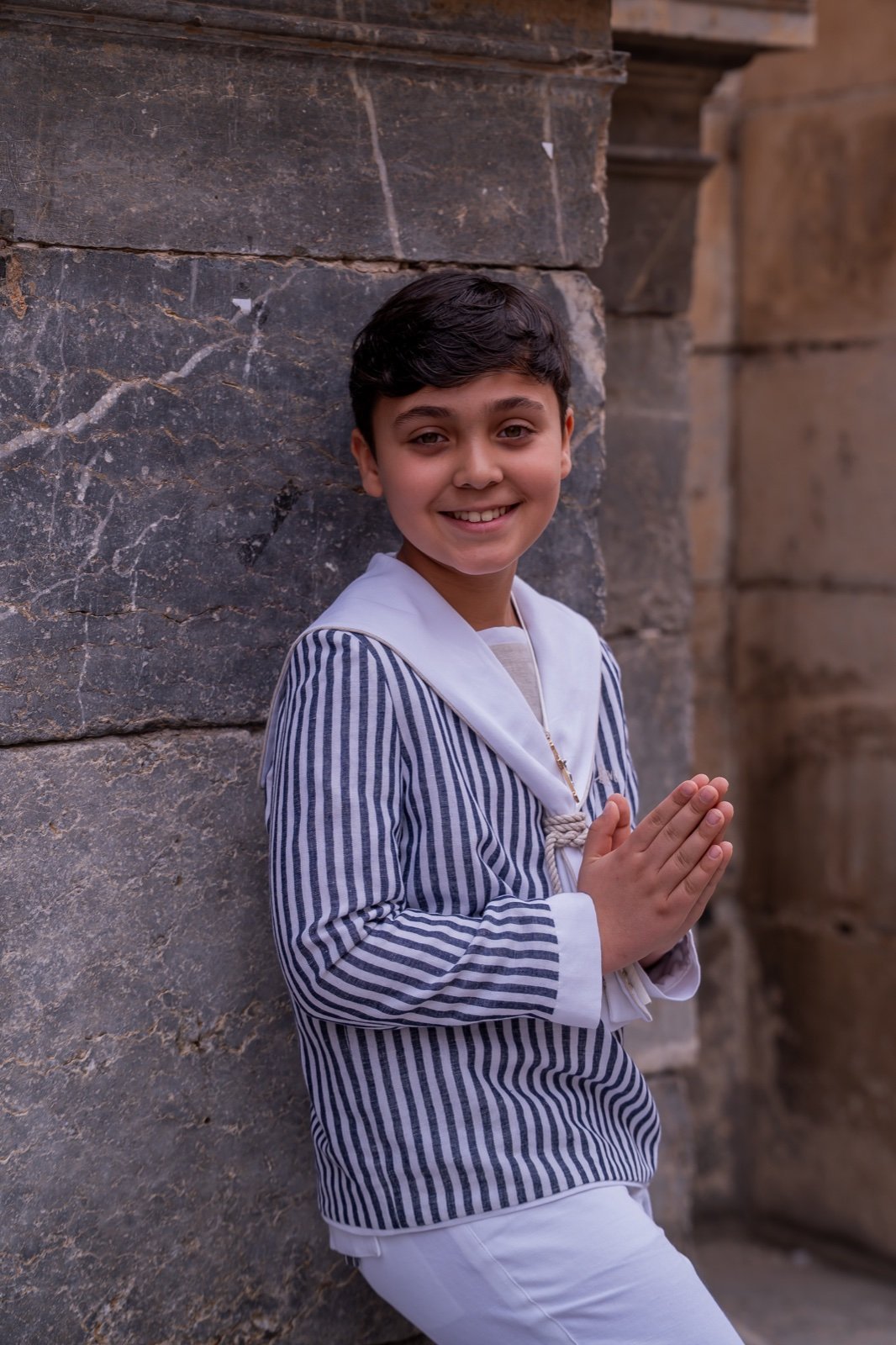 Niño con traje marinero sonriendo junto a un muro de piedra