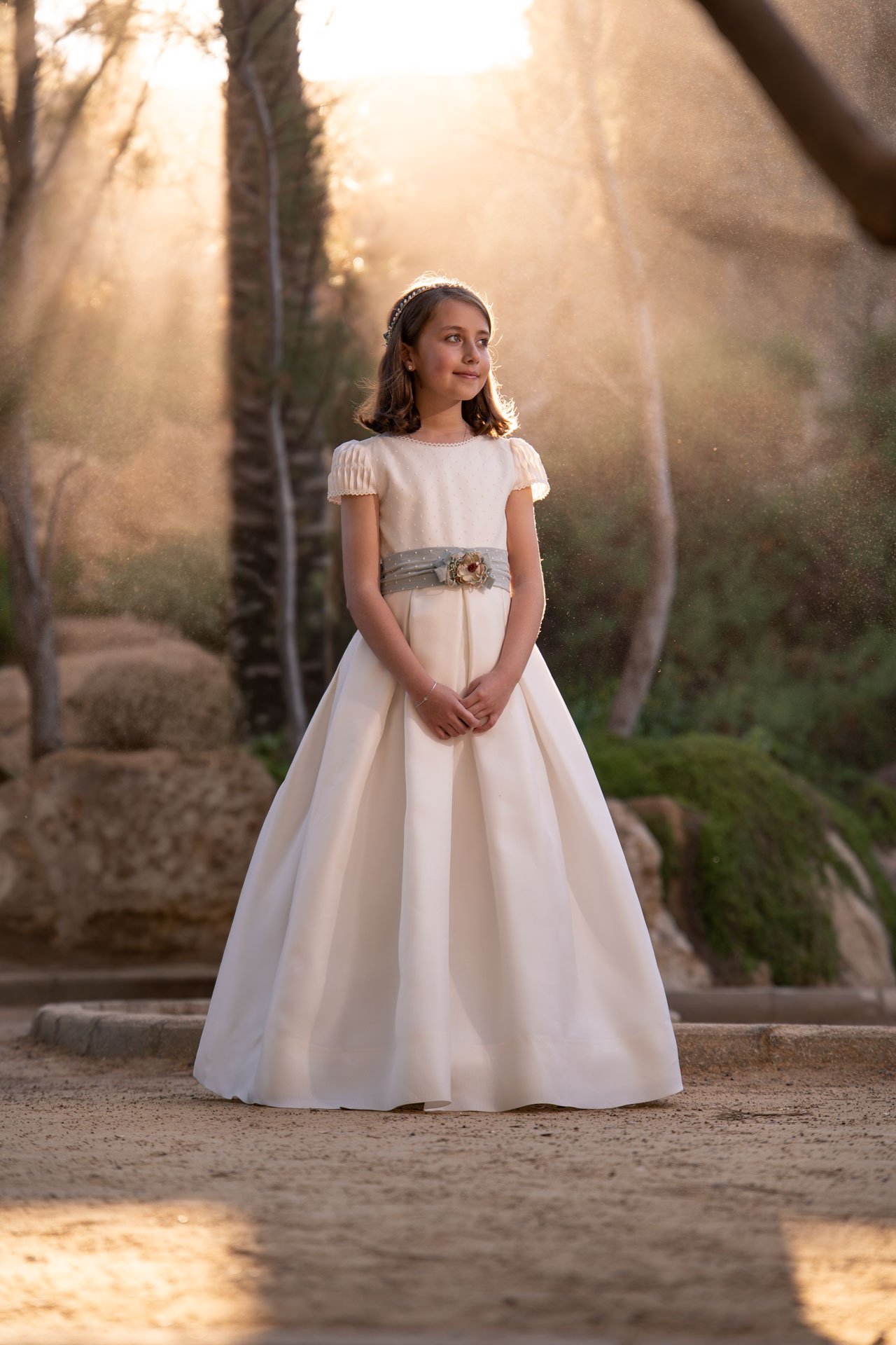 Una niña con vestido de comunión entre árboles iluminados por el sol.