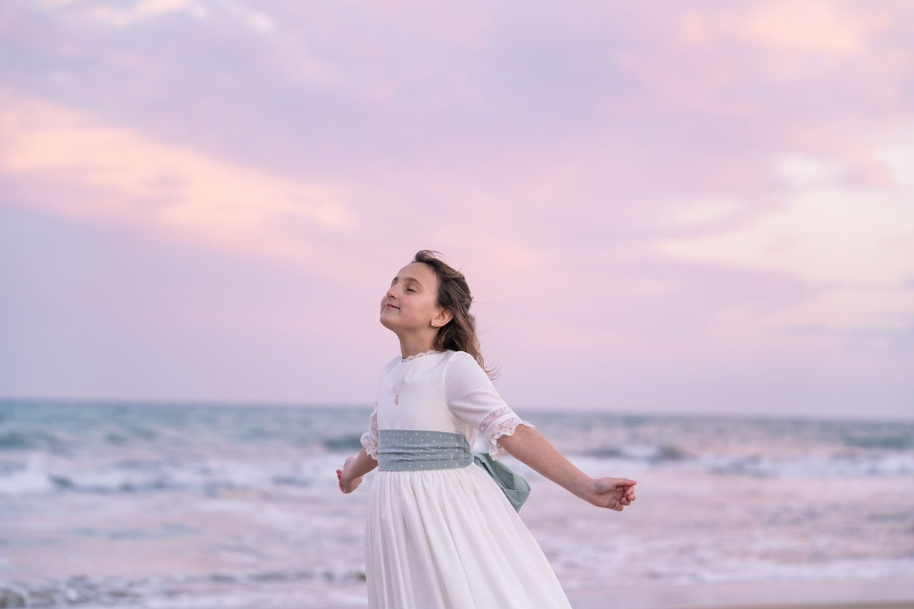 Una niña con vestido de comunión junto al mar bajo un cielo rosado.