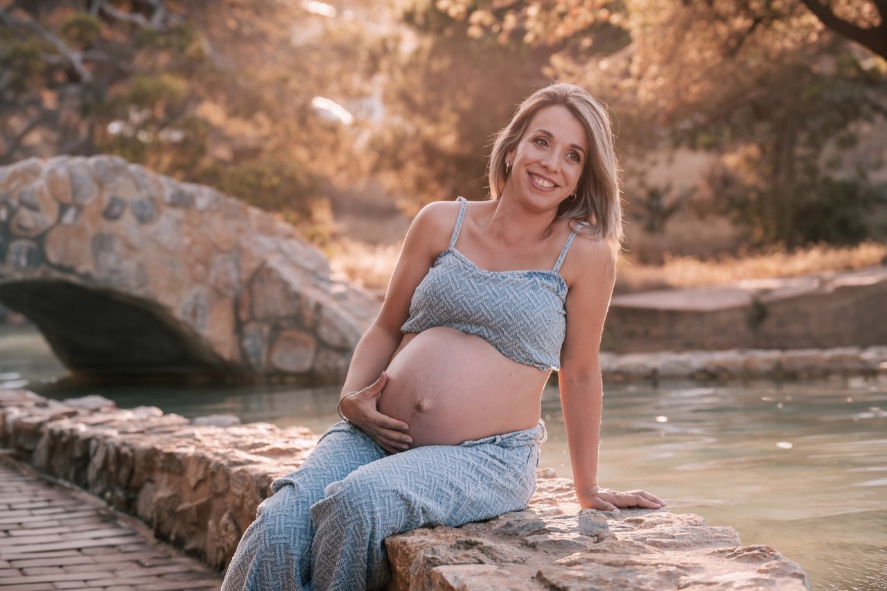 Mujer embarazada sentada junto al agua y un puente de piedra al atardecer